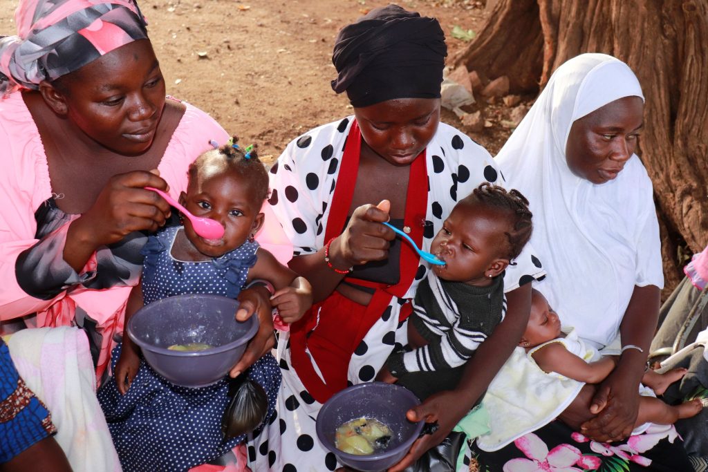 Two mothers, each balancing young children on their laps, feed their children from bowls.
